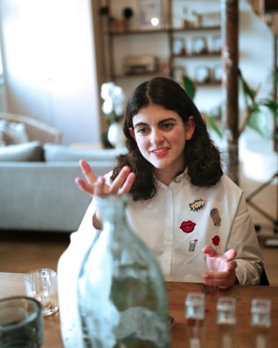 Jeune femme en chemise blanche animant une discussion sensorielle autour de flacons, dans un espace chaleureux et design.