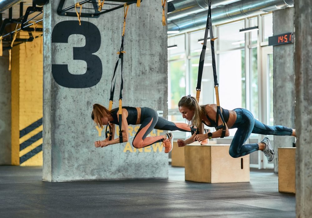 Two young athletic girls exercising with trx fitness straps at gym. Sport, partner workout, wellness and healthy lifestyle